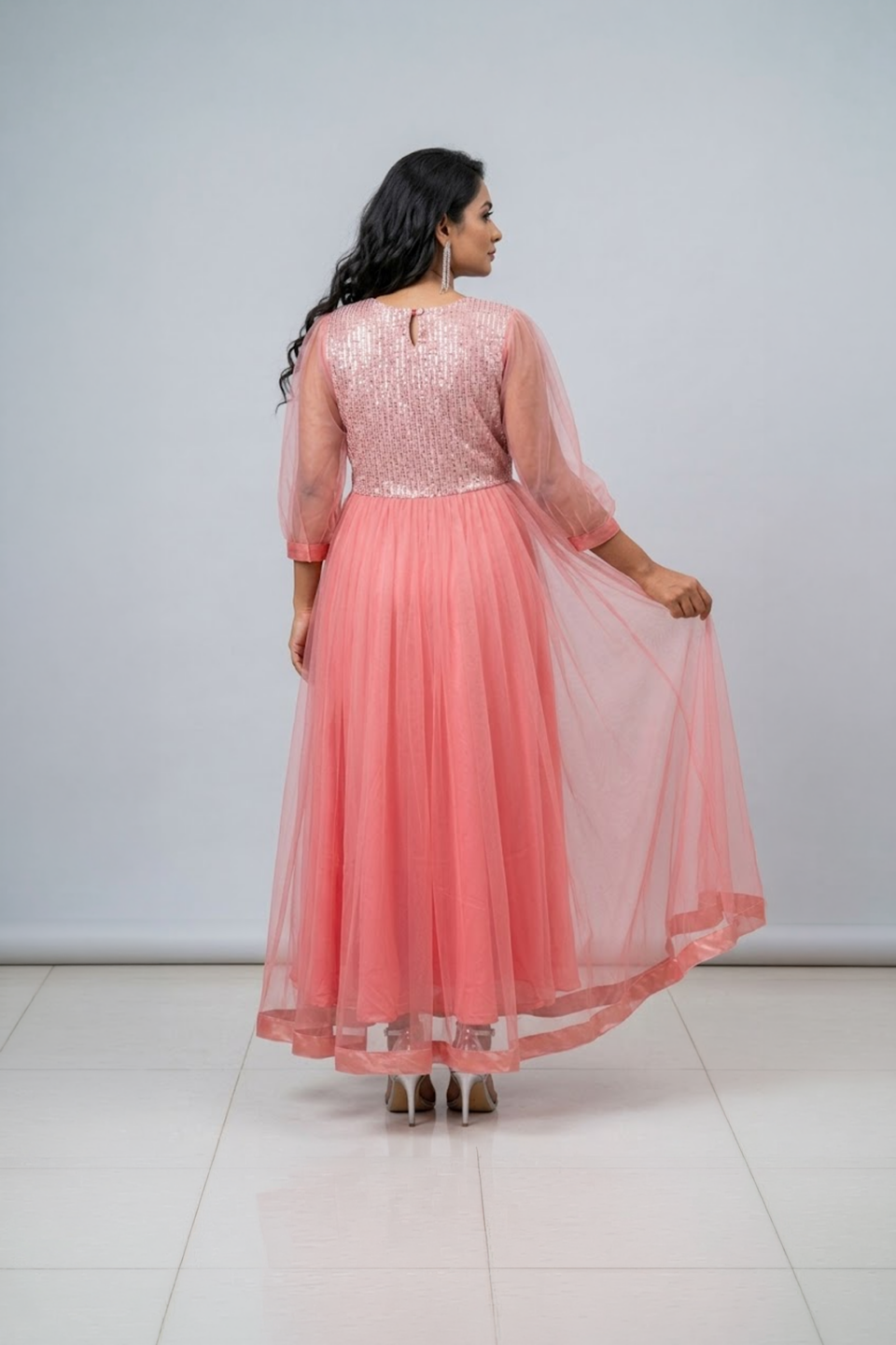 Woman wearing a coral pink shimmering tulle gown with a sequin bodice, square neckline and sheer three-quarter sleeves