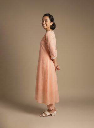 “Woman wearing a peach-orange embroidered kurta set with round neckline, keyhole detail, and matching pants, standing barefoot against a beige background.”