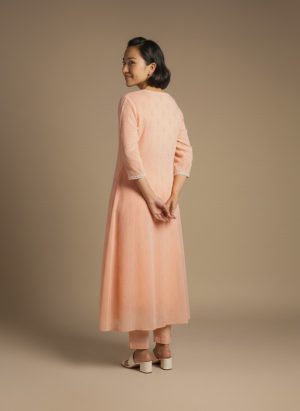 “Woman wearing a peach-orange embroidered kurta set with round neckline, keyhole detail, and matching pants, standing barefoot against a beige background.”