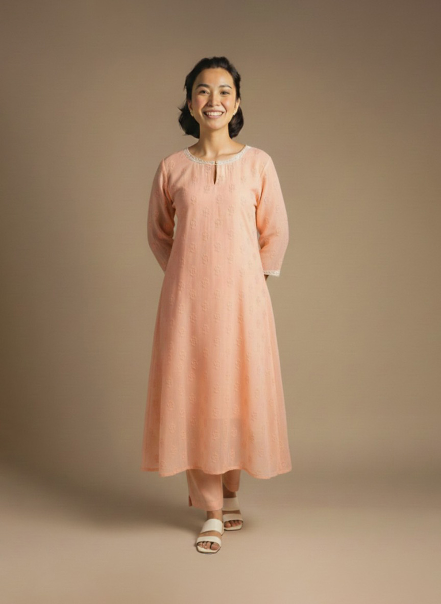“Woman wearing a peach-orange embroidered kurta set with round neckline, keyhole detail, and matching pants, standing barefoot against a beige background.”