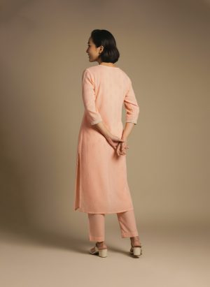 “Woman wearing a peach-orange embroidered kurta set with round neckline, keyhole detail, and matching pants, standing barefoot against a beige background.”