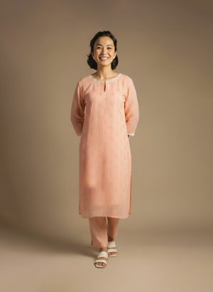 “Woman wearing a peach-orange embroidered kurta set with round neckline, keyhole detail, and matching pants, standing barefoot against a beige background.”
