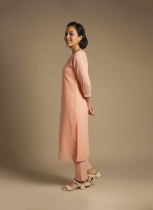 “Woman wearing a peach-orange embroidered kurta set with round neckline, keyhole detail, and matching pants, standing barefoot against a beige background.”