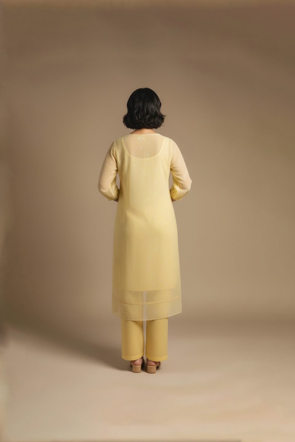 “Woman wearing a soft yellow knee-length dress with sheer long sleeves, subtle embroidery, and a relaxed fit, standing against a beige background and smiling.”