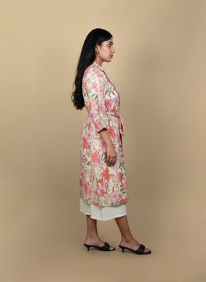 "Full view of a woman wearing a floral printed tie-waist long shrug layered over a white midi dress, posing against a beige background"