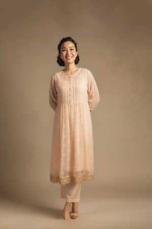 “Woman wearing a peach Kota embroidered kurta set with gold lace border and sheer full sleeves, standing against a beige background.”
