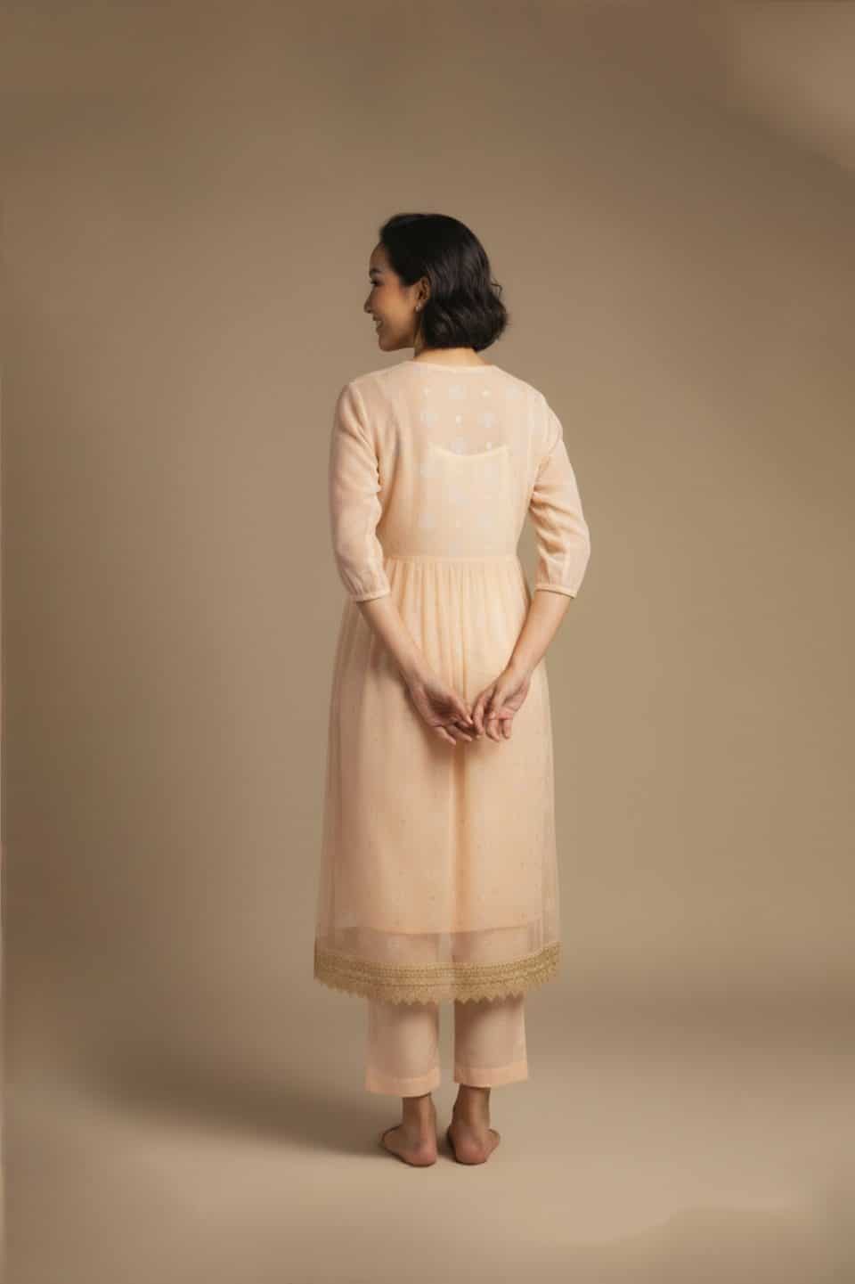 “Woman wearing a peach Kota embroidered kurta set with gold lace border and sheer full sleeves, standing against a beige background.”