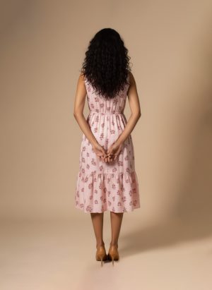 “Woman wearing a sleeveless pink cotton floral midi dress with a gathered waist and tiered hem, standing against a beige background.”