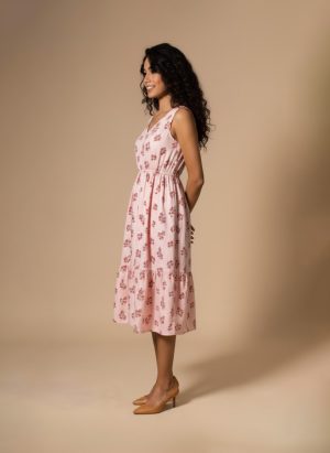 “Woman wearing a sleeveless pink cotton floral midi dress with a gathered waist and tiered hem, standing against a beige background.”
