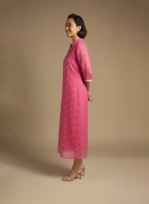 “Woman wearing a pink chikankari cotton kurta dress with white floral embroidery, V-notch neckline, and 3/4 sleeves standing against a beige background.”