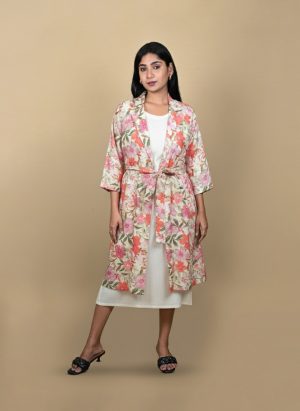 "Full view of a woman wearing a floral printed tie-waist long shrug layered over a white midi dress, posing against a beige background"