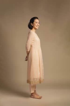 “Woman wearing a peach Kota embroidered kurta set with gold lace border and sheer full sleeves, standing against a beige background.”