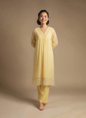 “Woman wearing a soft yellow knee-length dress with sheer long sleeves, subtle embroidery, and a relaxed fit, standing against a beige background and smiling.”