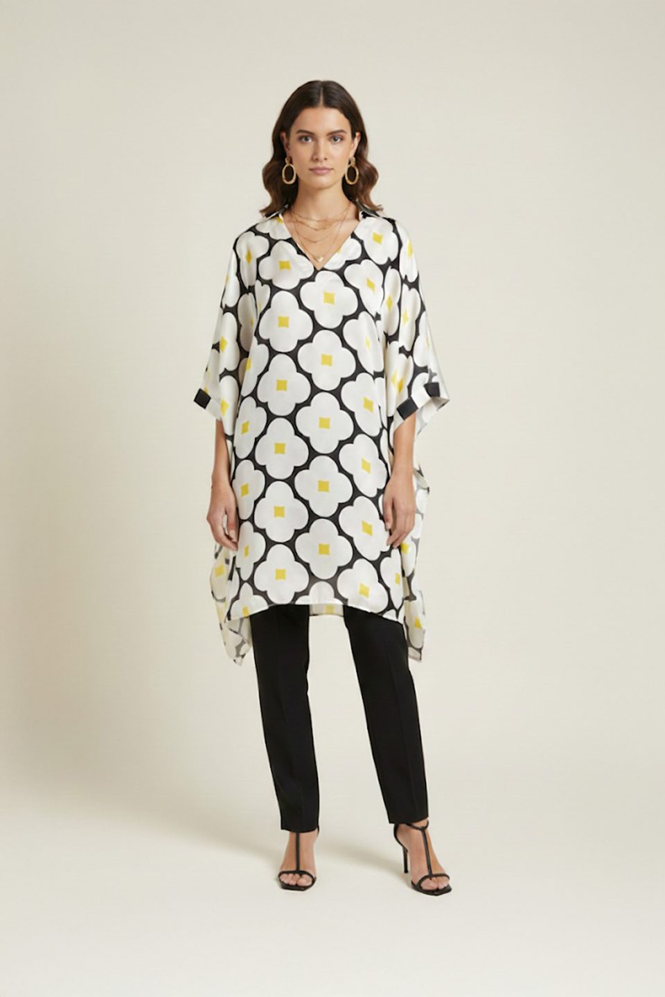 Woman wearing a black and white oversized floral printed tunic with yellow accents, featuring a relaxed silhouette and V-neckline, paired with slim black trousers and heels.