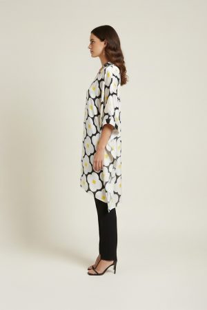 Woman wearing a black and white oversized floral printed tunic with yellow accents, featuring a relaxed silhouette and V-neckline, paired with slim black trousers and heels.