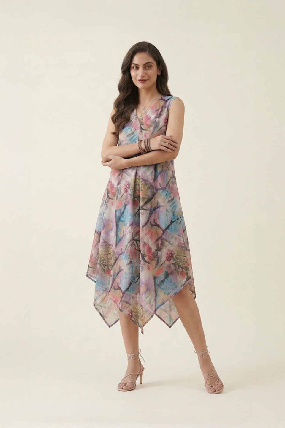 Woman wearing a pastel abstract floral printed sleeveless midi dress with an asymmetrical handkerchief hem, styled with strappy heels against a neutral background.